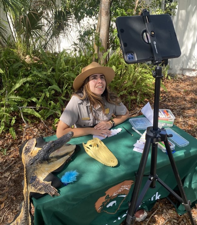 A park ranger sits outdoors at a table, facing a device on a tripod. There are alligator skulls and art supplies on the table
