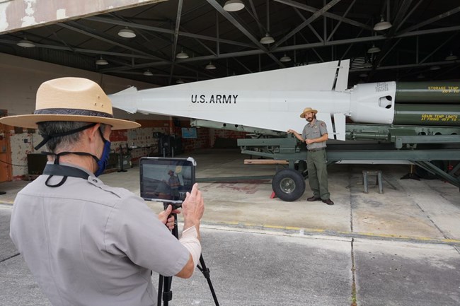 A ranger stands in front of a large white tipped missile which reads "US ARMY". Another ranger stands feet away with a camera broadcasting the missile and ranger.