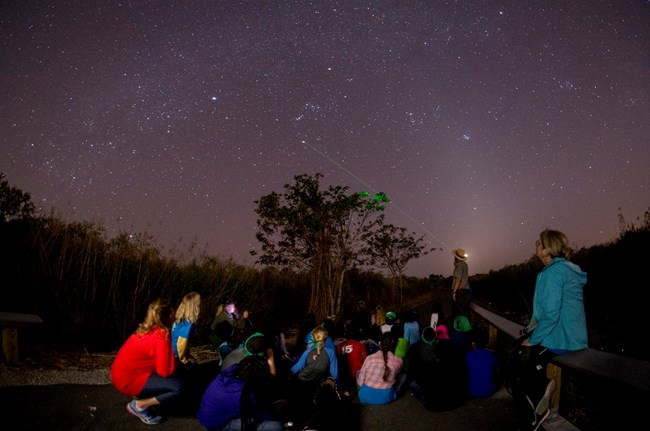 Everglades Astronomy Children sitting on paved trail at night. Park ranger points laser to the sky filled with stars.