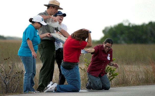 park ranger pours water on a board while adults standing and kneeling laugh