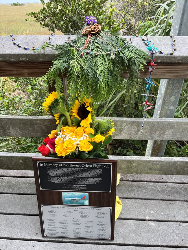 A plaque, vase of flowers, wreath of greenery and other mementos rest on the floor and railing of boardwalk overlook.