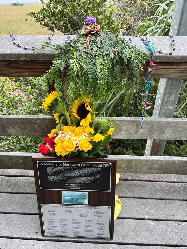A plaque, vase of flowers, wreath of greenery and other mementos rest on the floor and railing of boardwalk overlook.