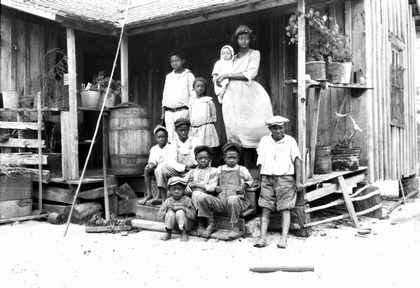 A black and white image of an African American woman and nine children gathered on the porch of a wooden home in the Everglades in 1929.