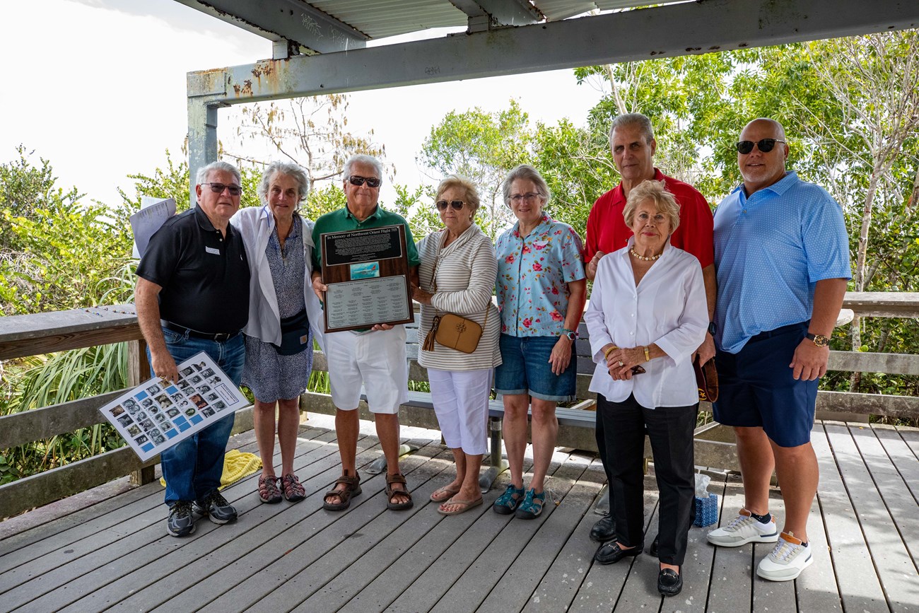 Eight men and women pose for a group photo with a plaque and laminated sheet on a covered deck