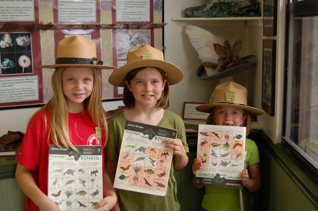 Three young girls wearing flat hats and holding Junior Ranger booklets pose for a photo