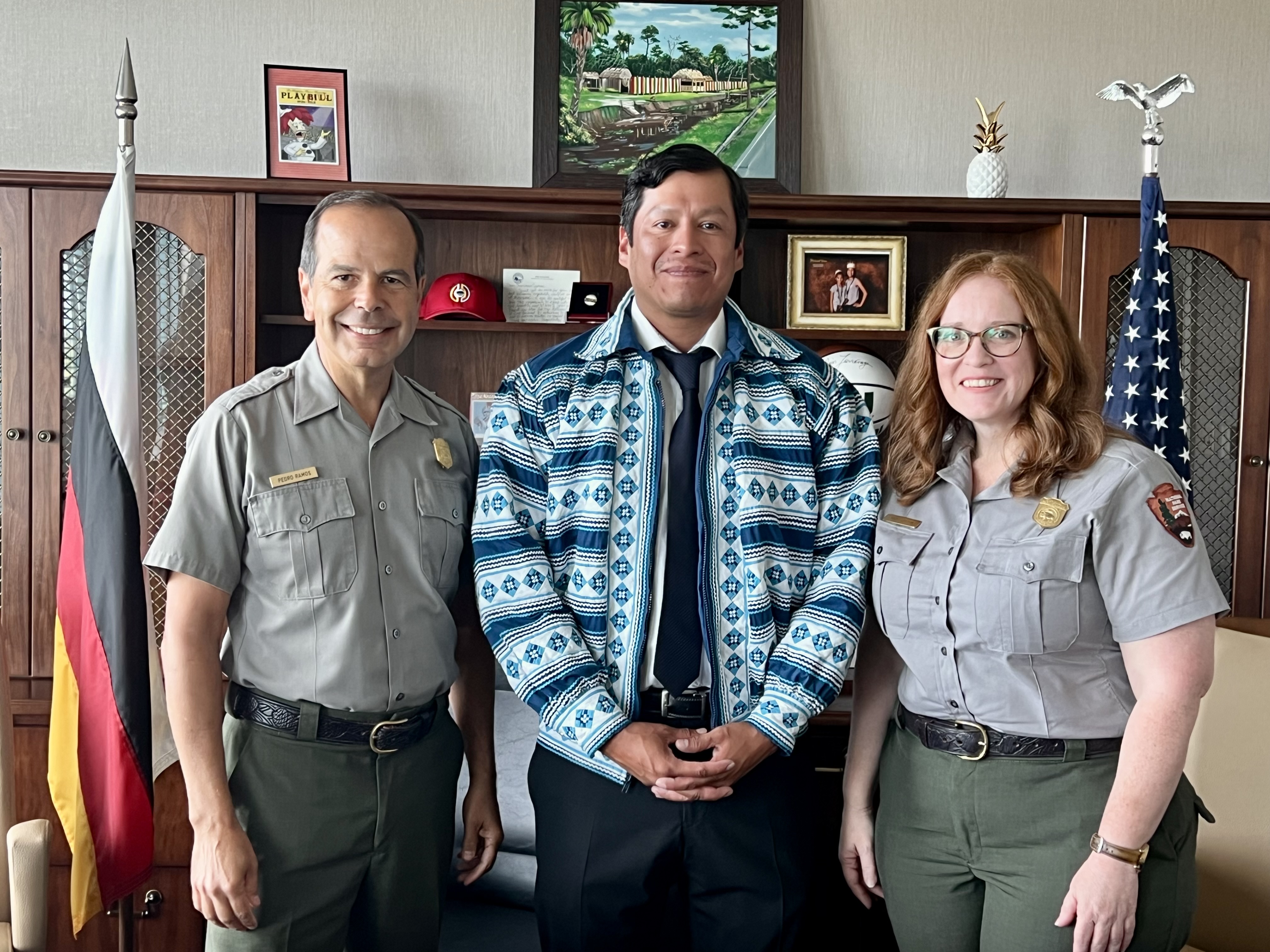 From left to right, a uniformed male park ranger, a Miccosukee Tribal official in traditional jacket, and a female uniformed park ranger stand in an office.