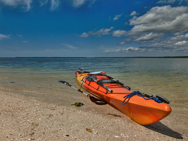 An orange and yellow kayak and paddle rest on a sandy beach with the ocean and a blue sky with clouds iin the background.