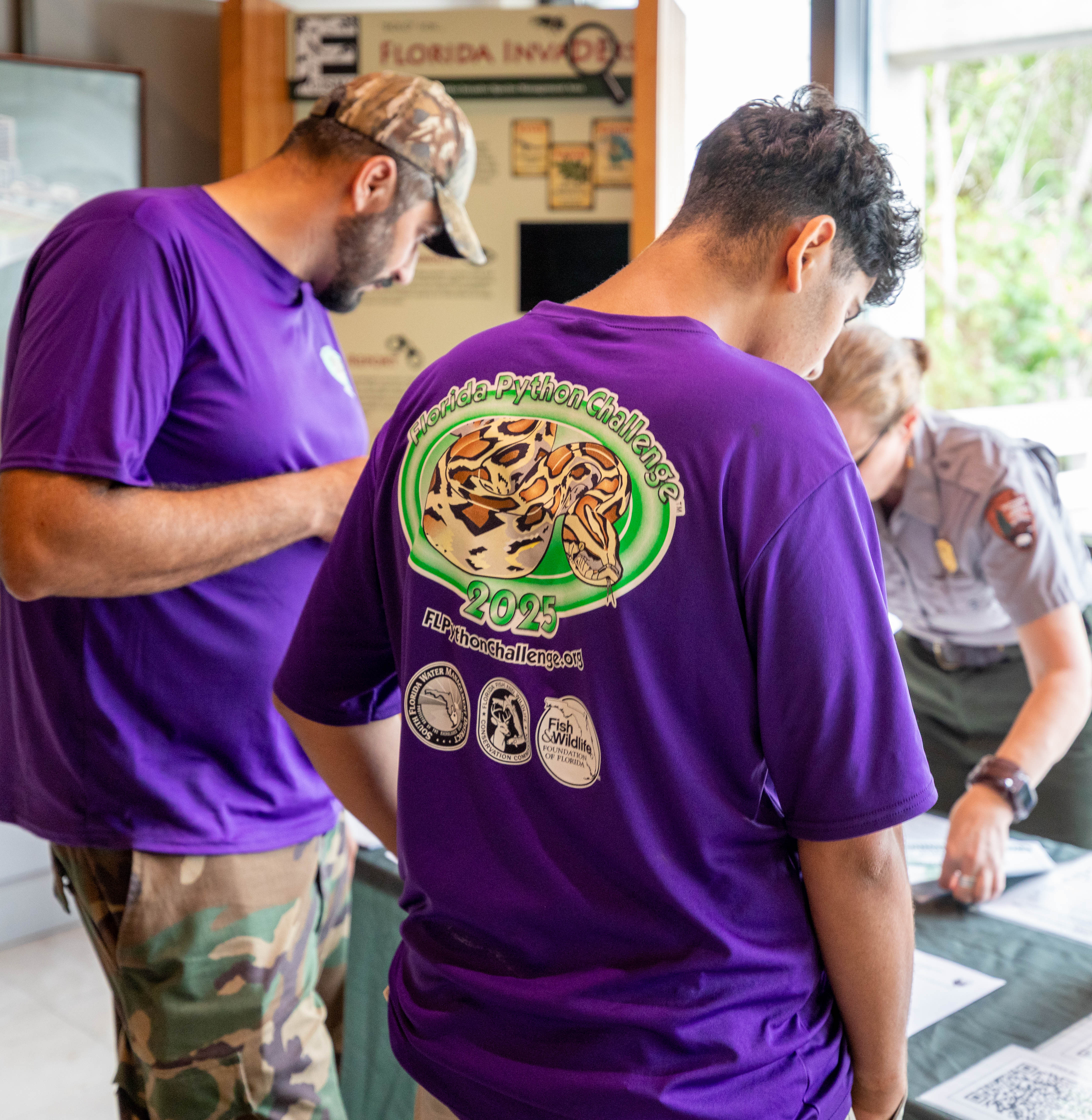 Two men in purple shirts work with a ranger checking them in at a table with a green table cloth.