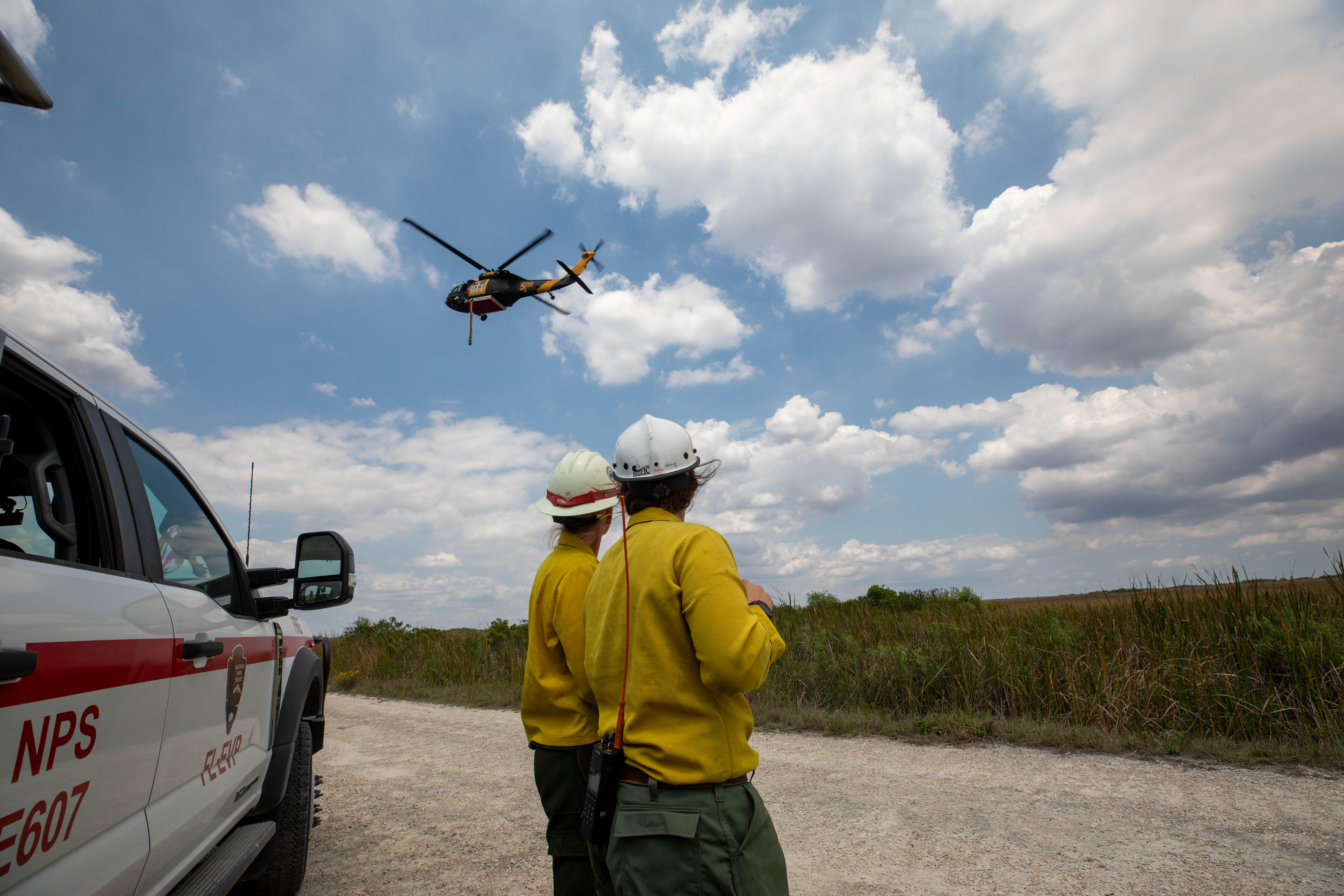 Two wildland firefighters in yellow shirts and white hard hats watch a helicopter while standing near a white truck with red stripe.