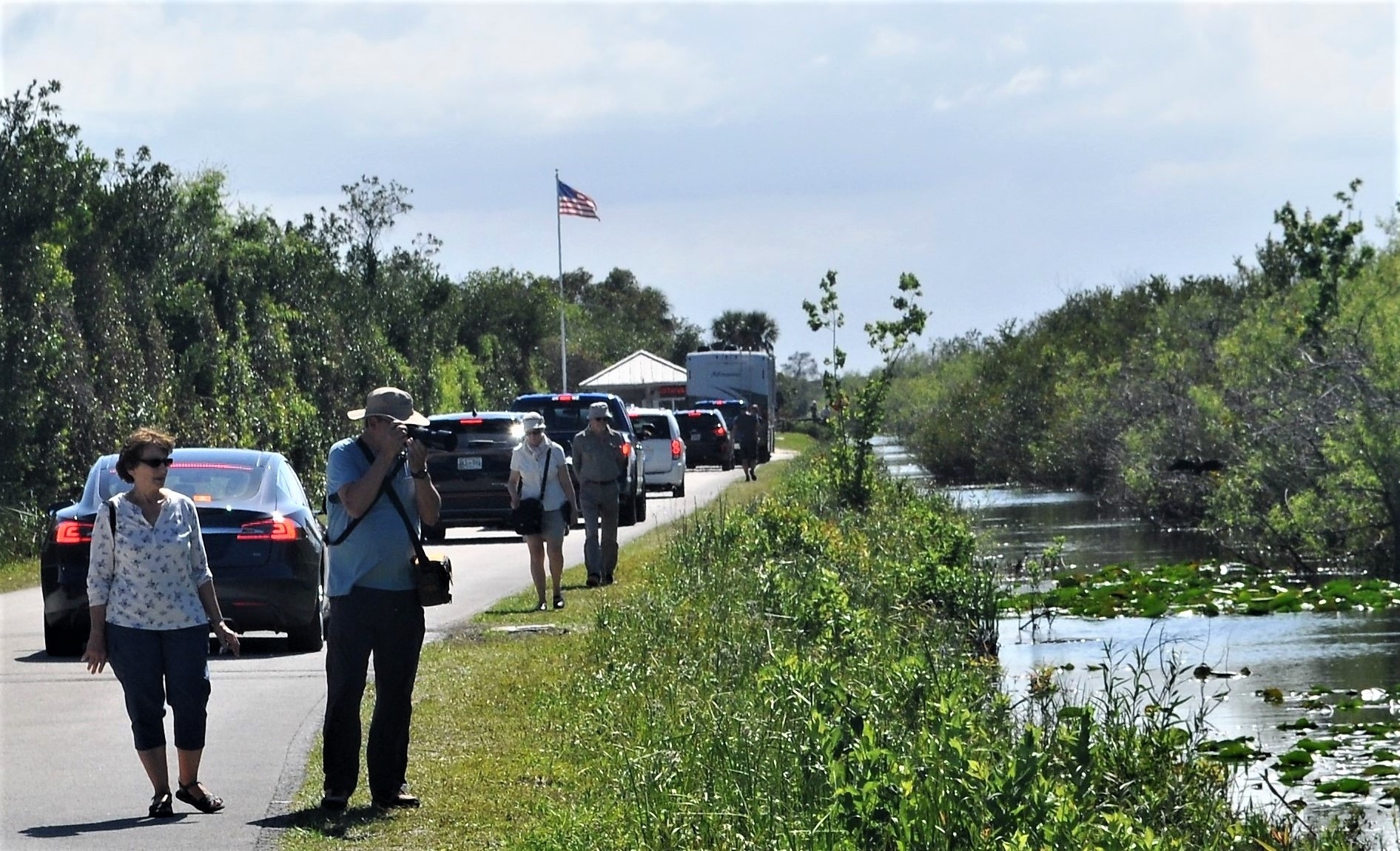 A line of cars waits at the entrance booth with an American flag flying above, while visitors stop along the road to take photographs of the adjacent canal with vegetation.