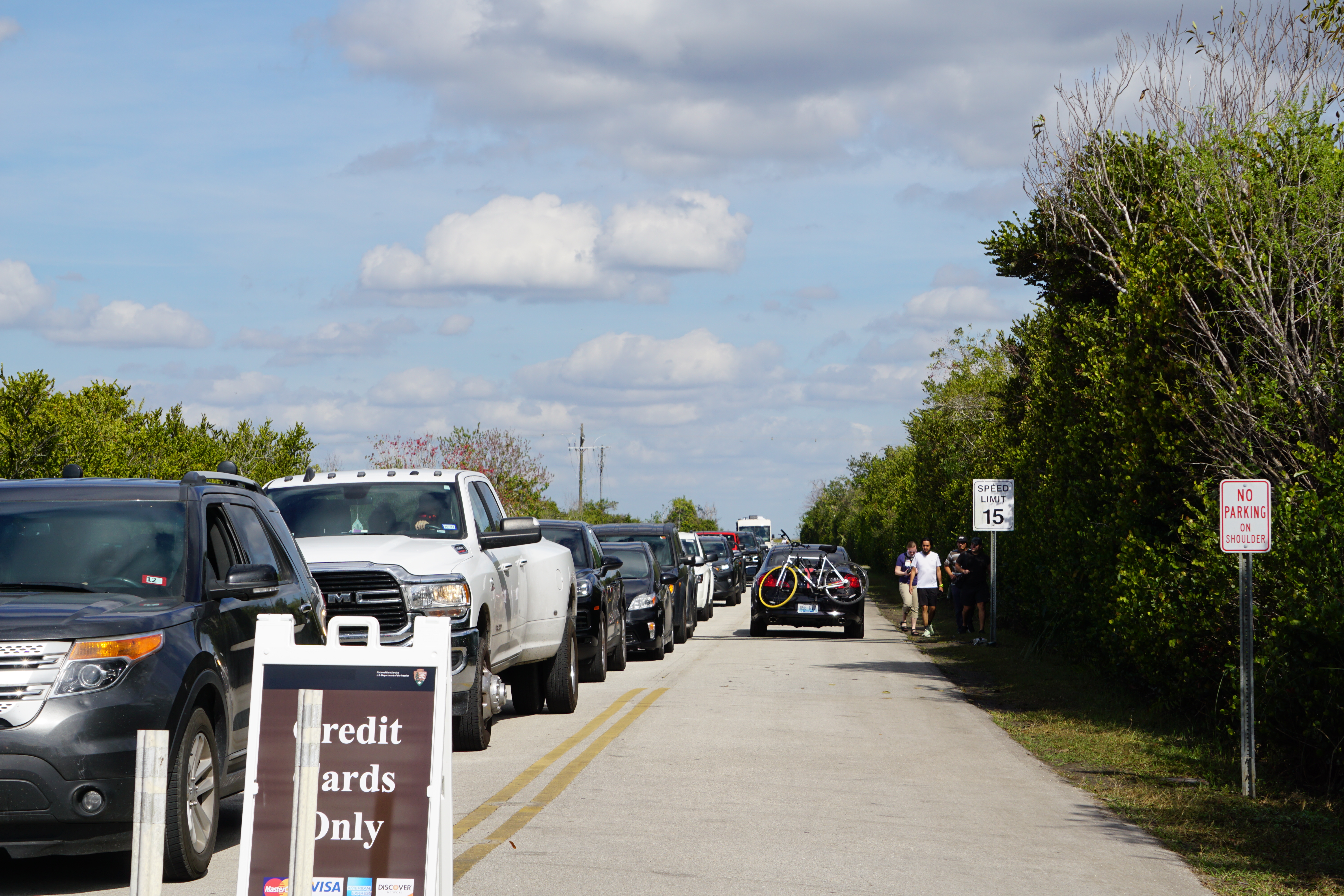 A line of vehicles facing forward in left lane while one car with a bicycle attached at the rear departs in the right lane. Visitors walk alongside the road. A sign in the foreground reads "Credit Cards only.