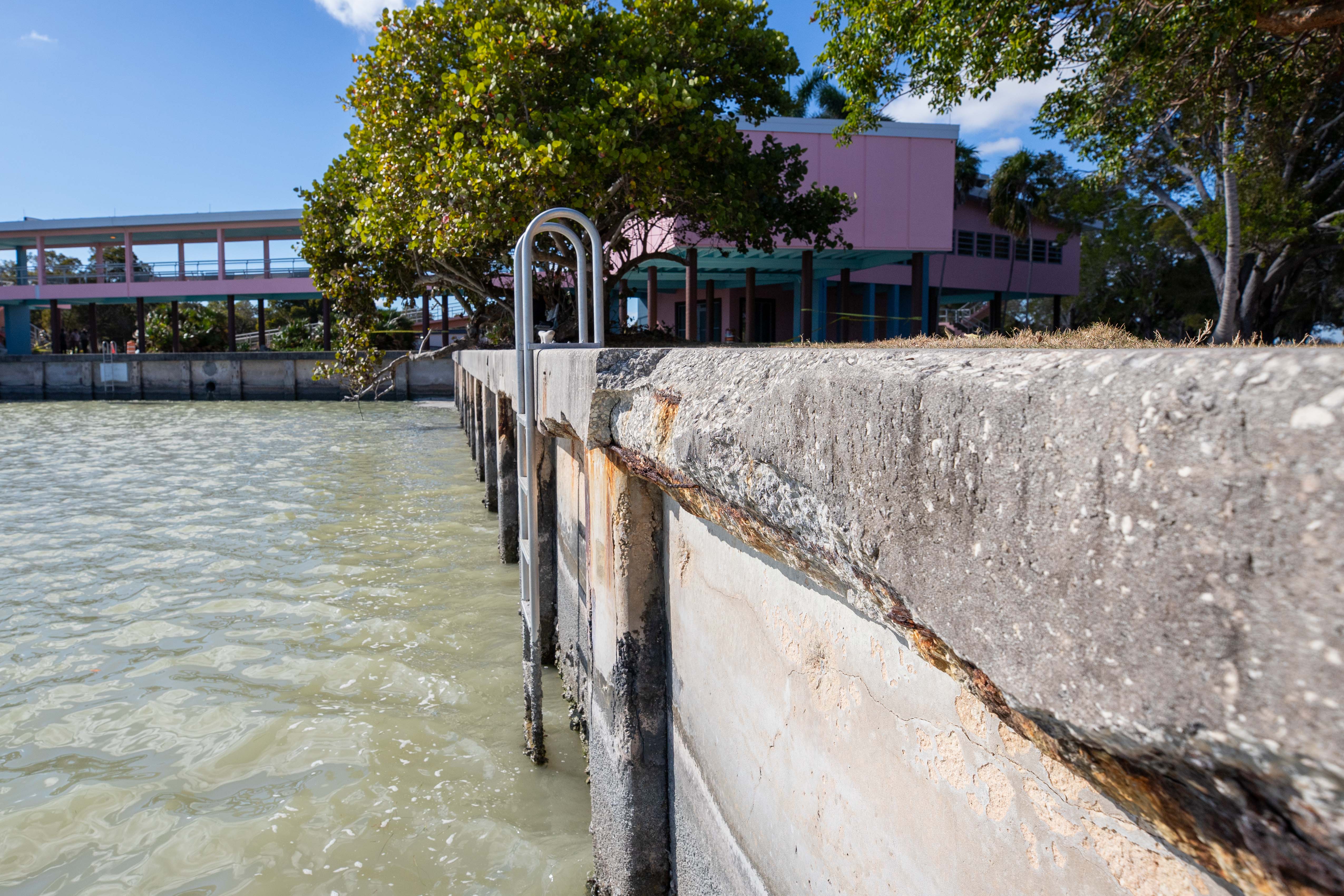 Concrete seawall with cracks and broken sections in front of a pink two-story building