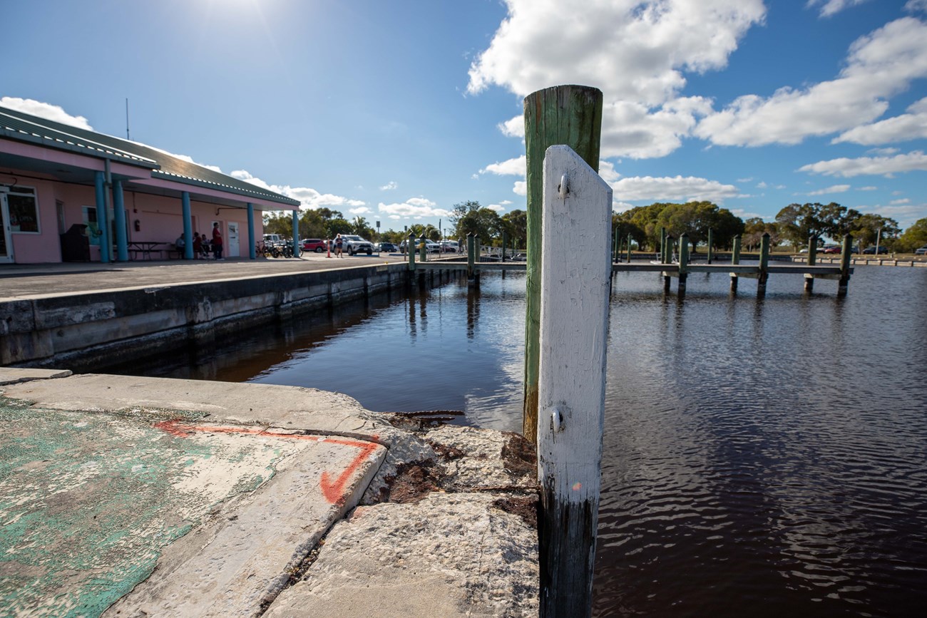 Broken edge of a concrete seawall next to a piling in front of water and a pink one-story building