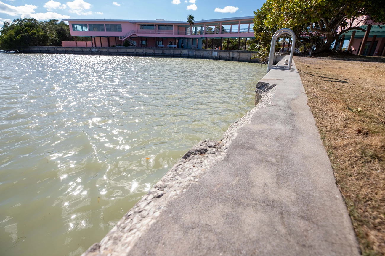 EVER_FlamingoSeaWall_14FEB2023 (7 of 16) A cement seawall with a longitudinal section missing lines a boat basin with the two-story pink building with open breezeway in the background.