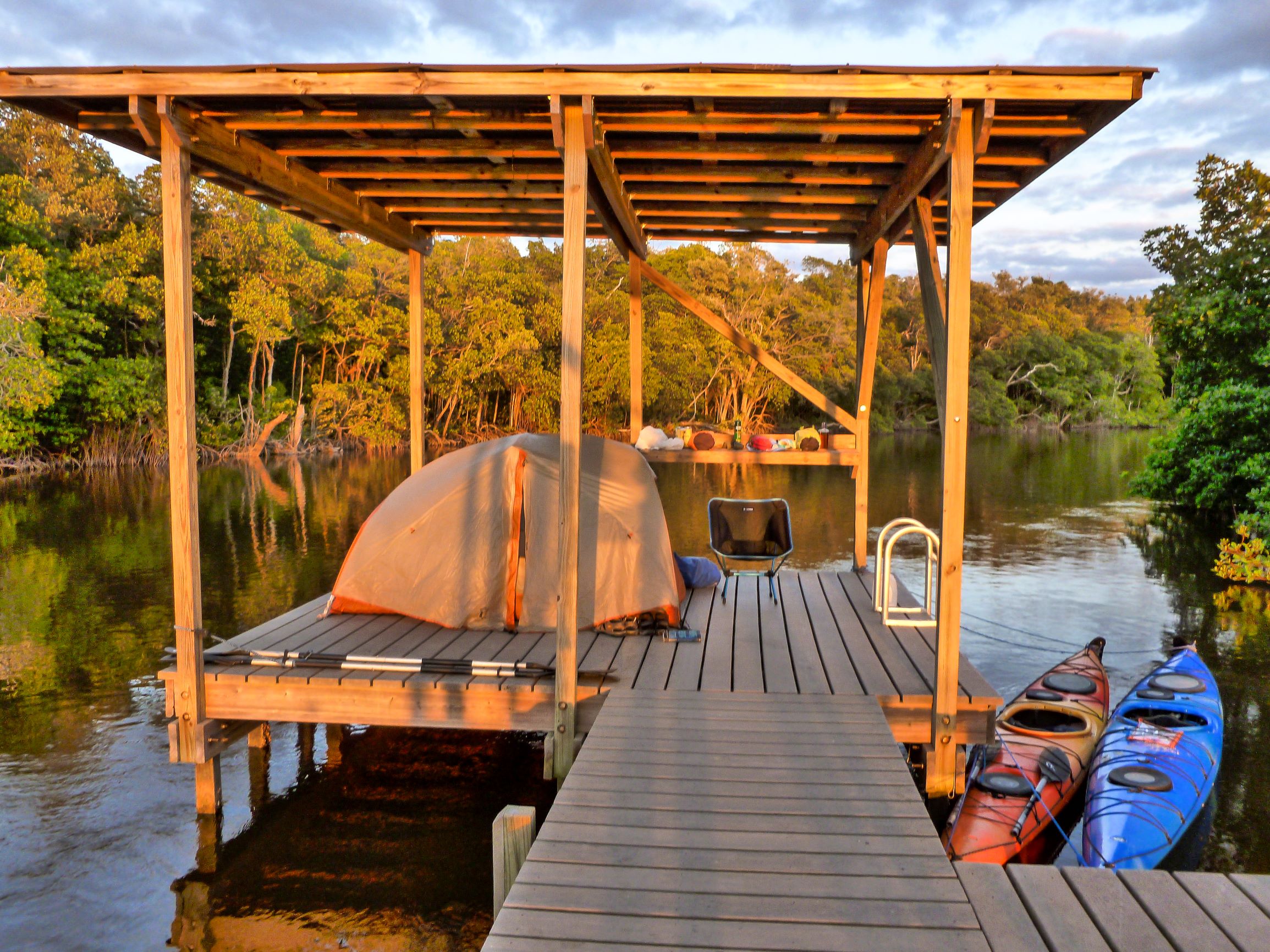 Elevated wooden camping platform with pitched tent and red and blue kayaks on a mangrove waterway.