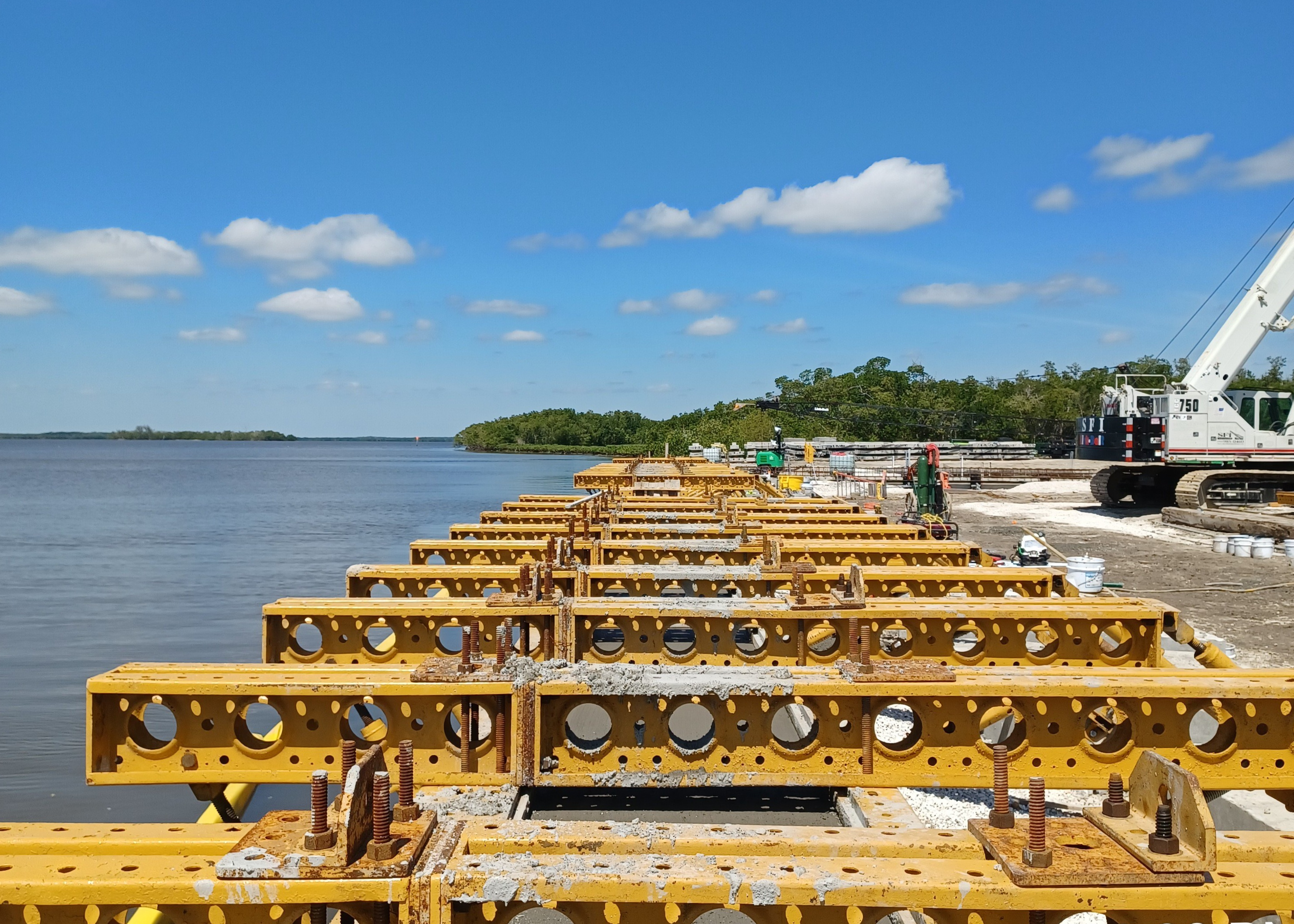 Large horizontal yellow forms are lined up in place for pouring concrete to construct a seawall. View of the open water and trees lining the distant shoreline.
