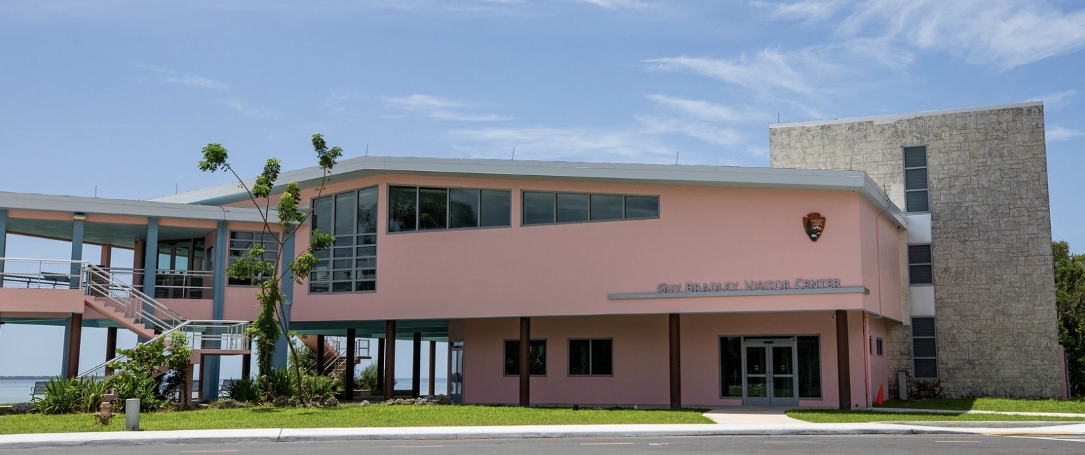 A bubble-gum-pink two-story building with partial view of breezeway and open water behind bears the NPS arrowhead logo and the words "Guy Bradley Visitor Center"