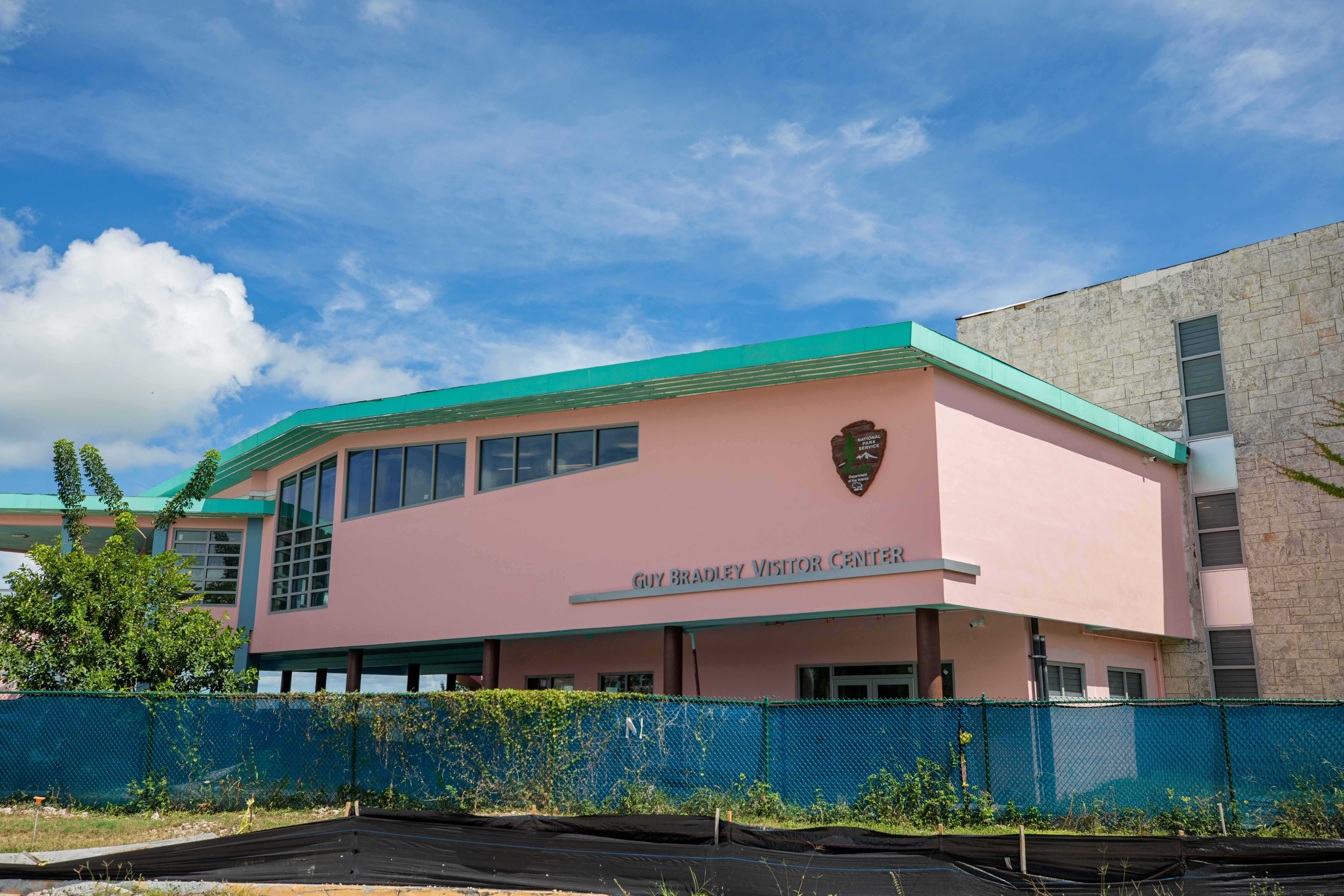 A pink building with turquoise roof is surrounded by blue construction fencing. National Park Service arrowhead and "Guy Bradley Visitor Center" are on the side of the building.