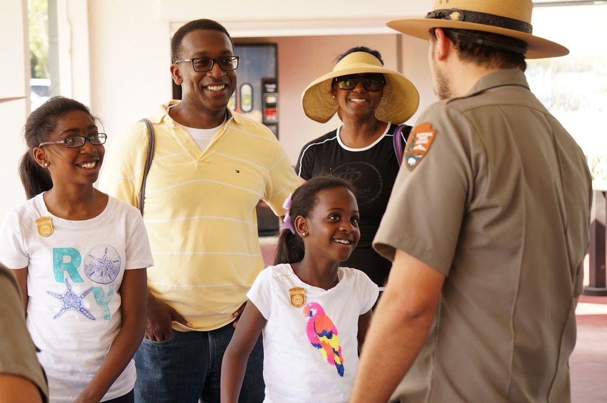 Two girls with Junior Ranger badges and their parents are smiling and facing a man in uniform with the National Park Service arrowhead patch on his left arm.