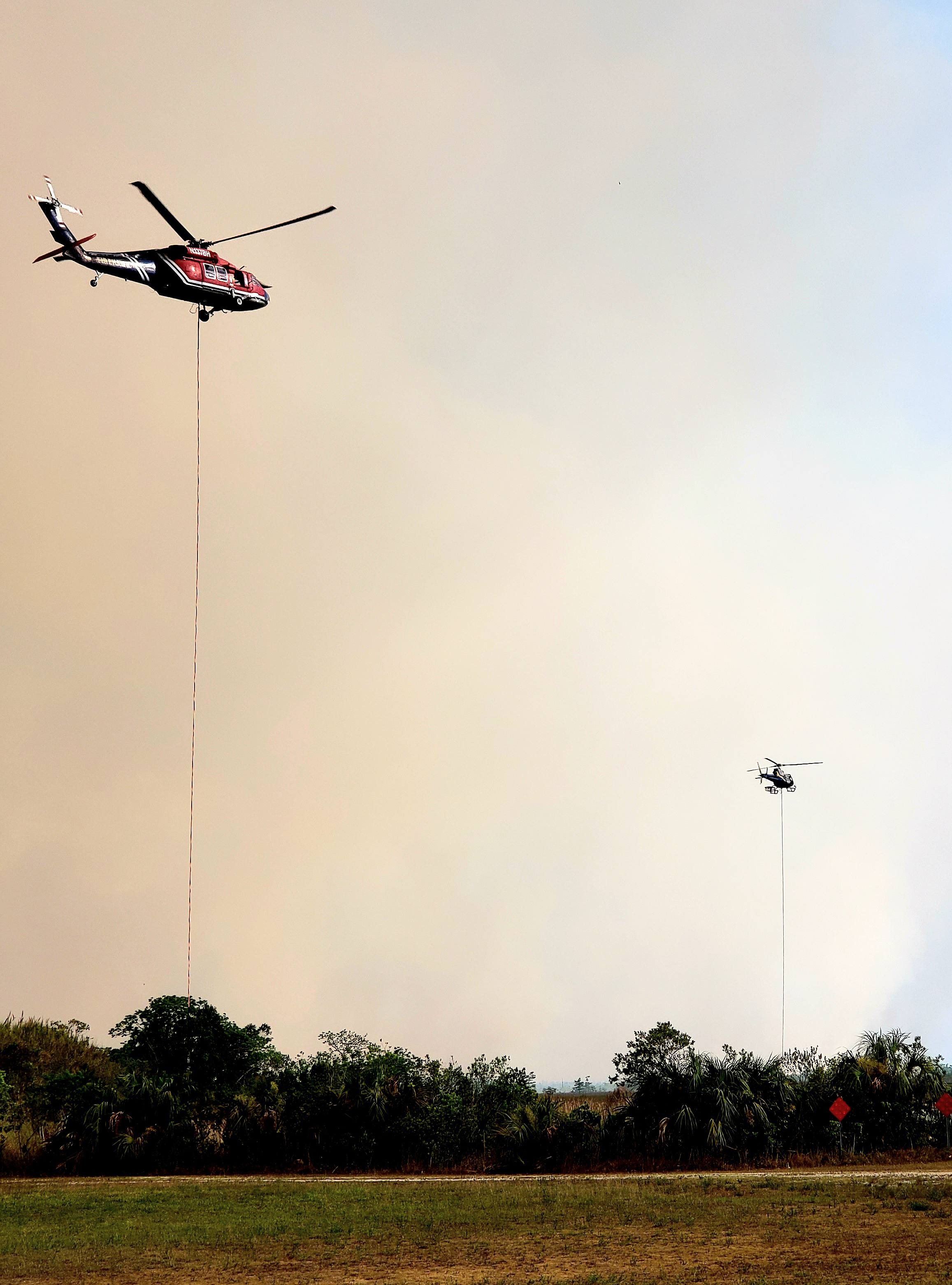 NPS Photo Moon Fish Wildfire Helicopters Two helicopters with lines hanging down fly above grass and shrubs with smoke clouds behind them.