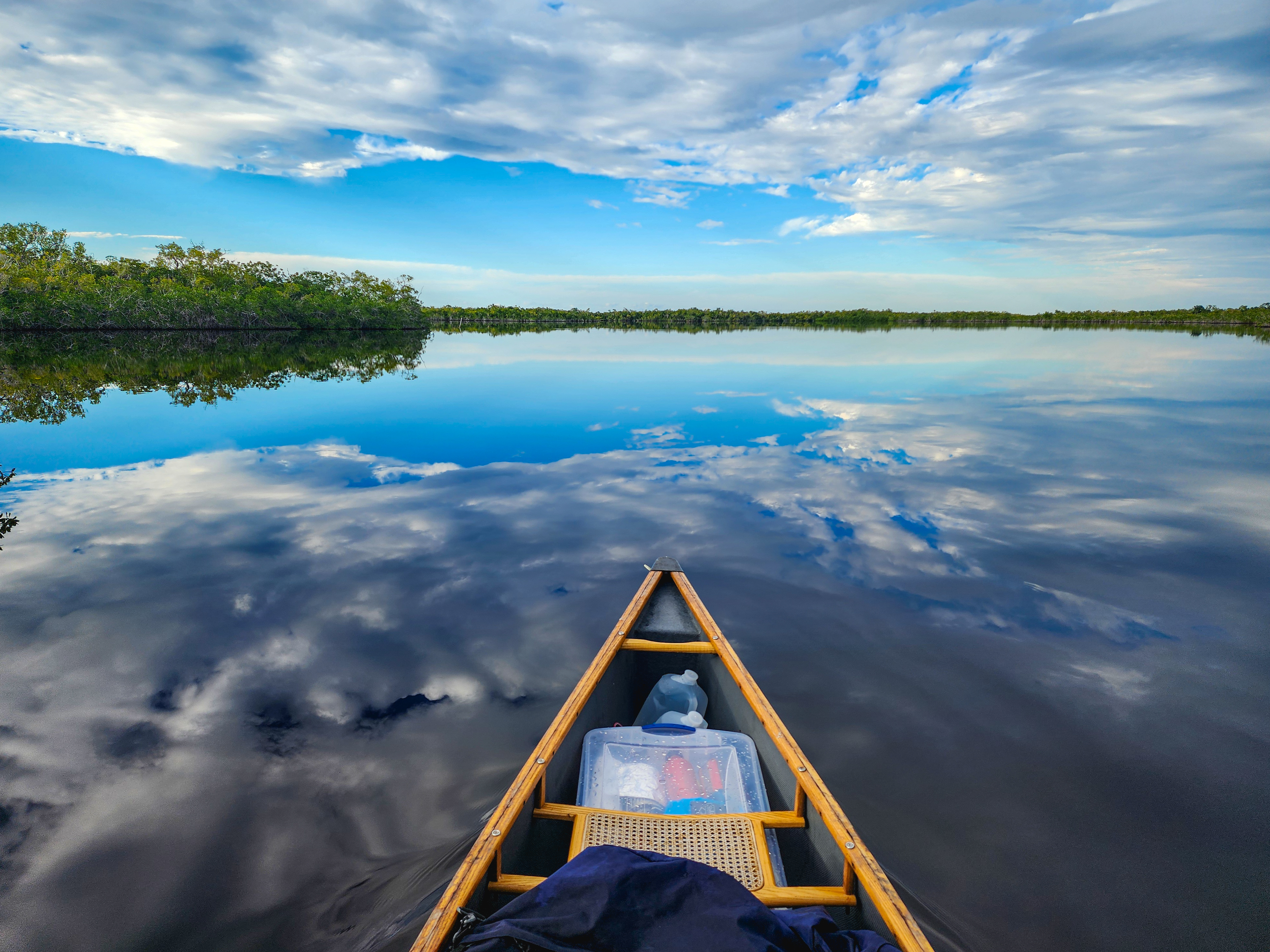 The bow of a canoe loaded with gear floats on open water with a perfect reflection of a blue sky with white clouds. The surrounding shorelines are lined with mangrove trees in the distance,