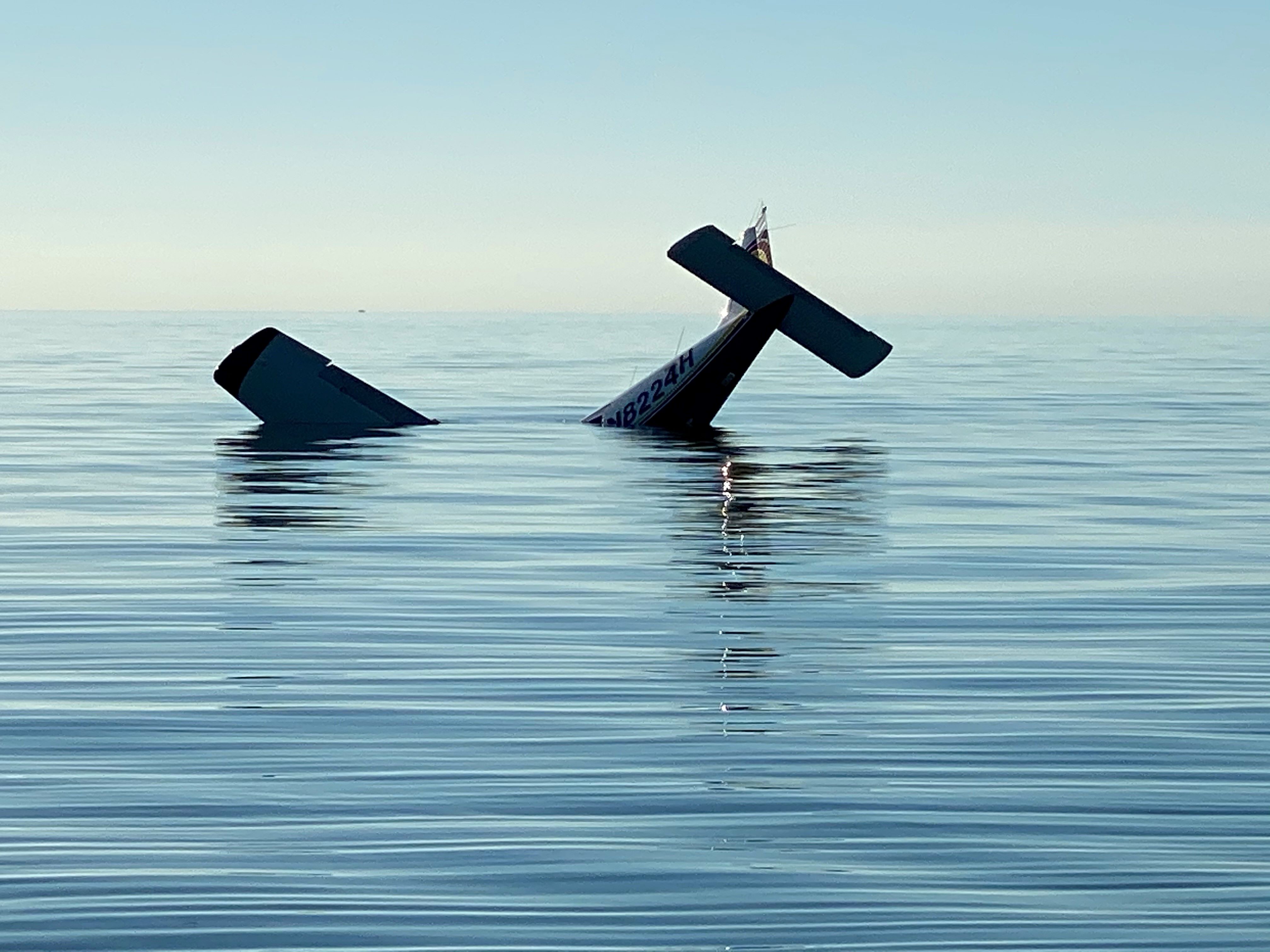 The tail and one wing of a submerged airplane protrude from calm blue water under clear blue sky.