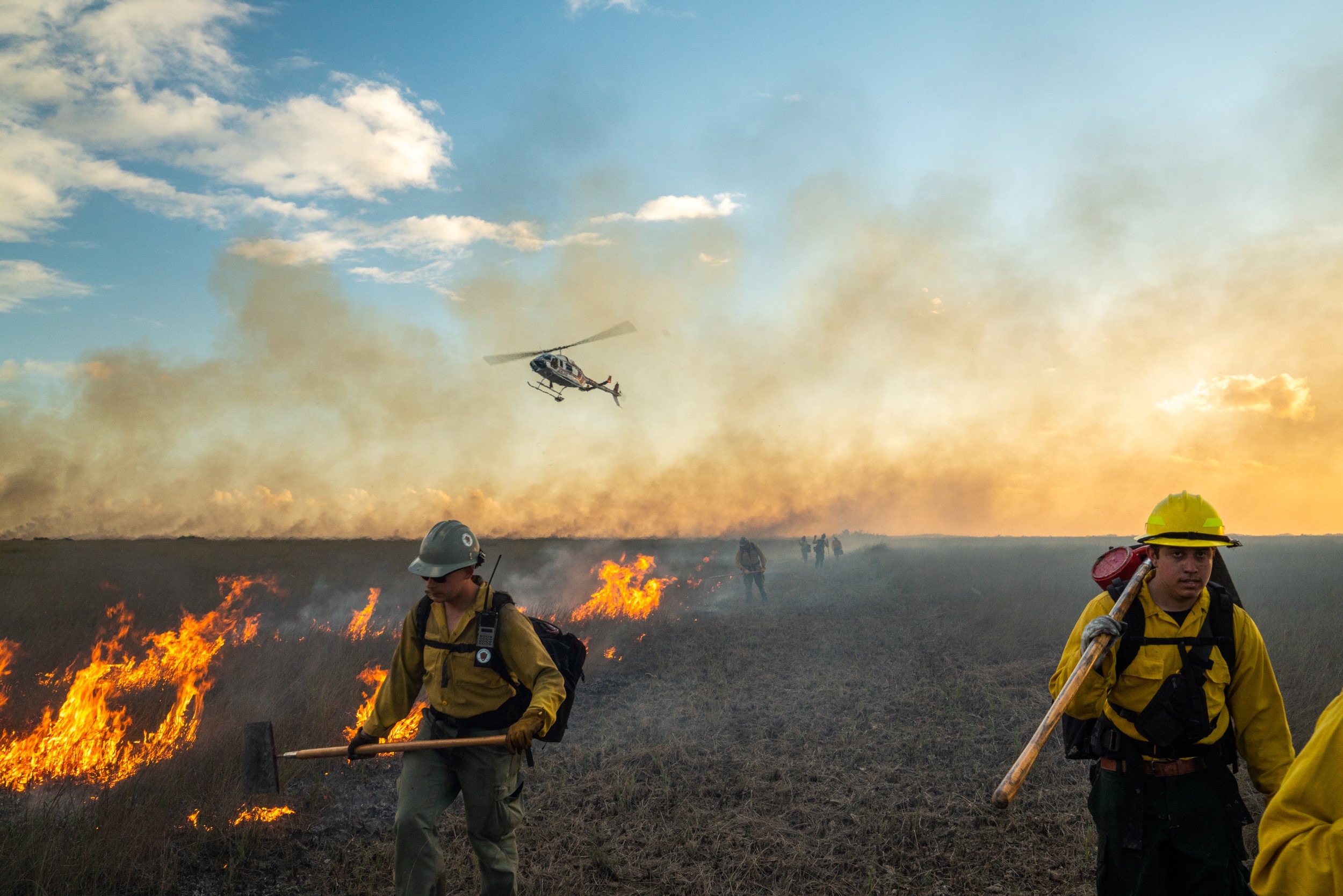 Two firefighters wearing safety gear and carrying tools walk away from a line of flames as a helicopter hovers close to the ground in the distance.
