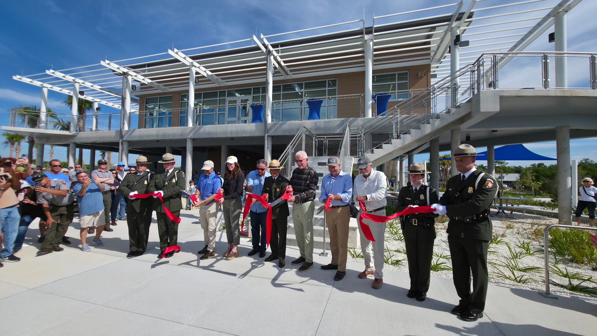 Image shows a line of individuals cutting a red ribbon. There is a building behind them and onlookers on either side of them. The sky behind them is blue.