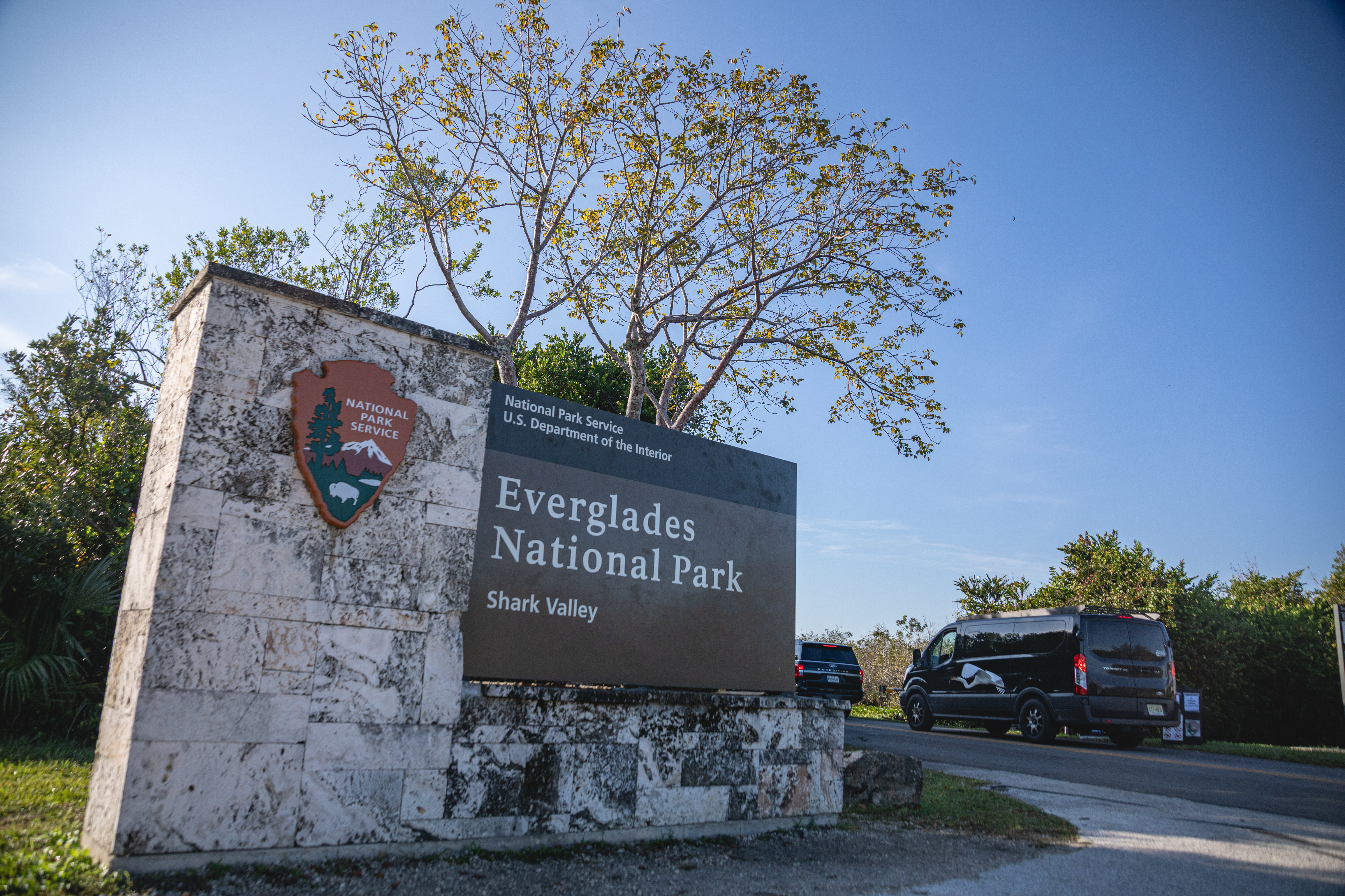 Images shows picture of park entrance sign that reads Everglades National Park. There are vehicles driving into the park.