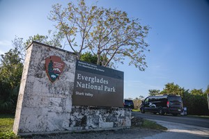 Images shows picture of park entrance sign that reads Everglades National Park. There are vehicles driving into the park.