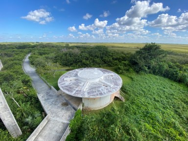 An aerial view of a paved pathway and the round-roofed building surrounded by greenery and view of the Everglades in all directions.