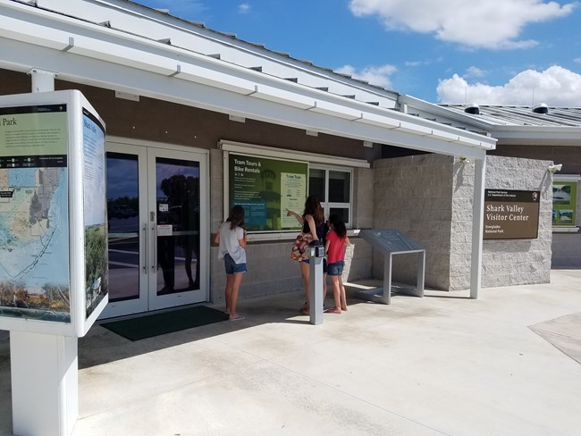 Exterior of the Shark Valley Visitor Center, with three park visitors viewing informational sign about tram tours and bike rentals
