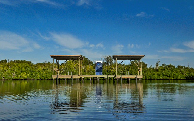 A raised platform above water, with trees and a blue sky in the background. There is a portable toilet on the platform and 2 covered camping areas.