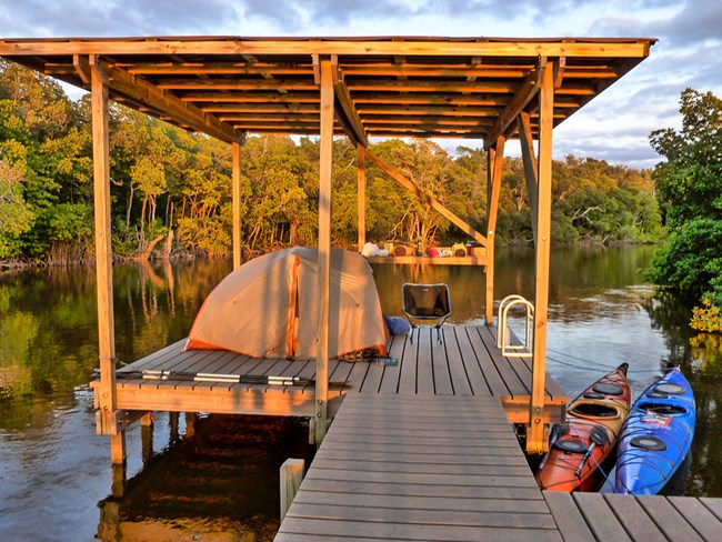 Two kayaks floating in the water tied to a wooden platform with a roof. On the platform are a tent and chairs.
