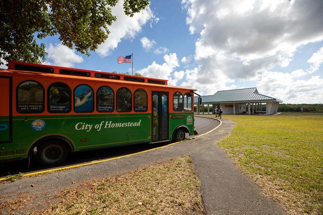 A green and orange trolley pulls up on a road with a flagpole and a building with a green roof in the background.