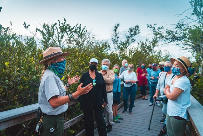 A Park Ranger speaks to a crowd of visitors on a boardwalk. Everyone is wearing a face covering.