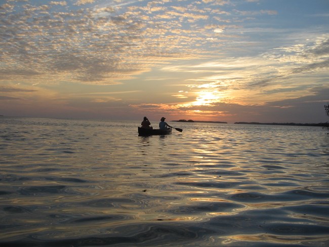 Two people in a canoe paddle over calm water while the sun sets