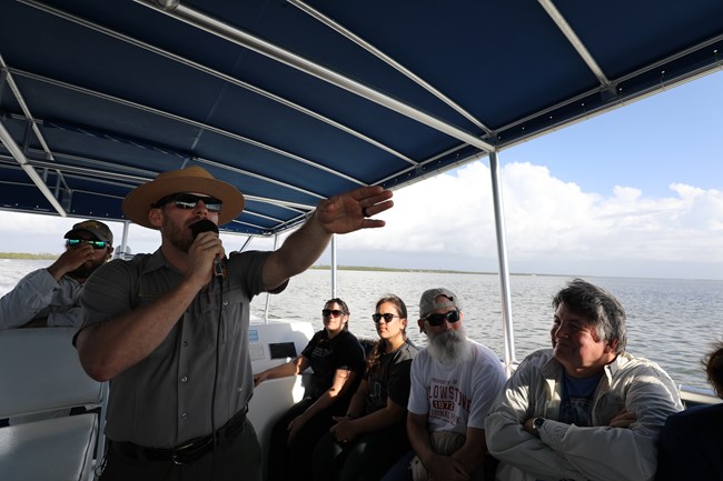 A male Park Ranger wearing sunglasses and flat hat holds a microphone in one hand and points with the other. He is aboard a boat and there are four visitors watching him.
