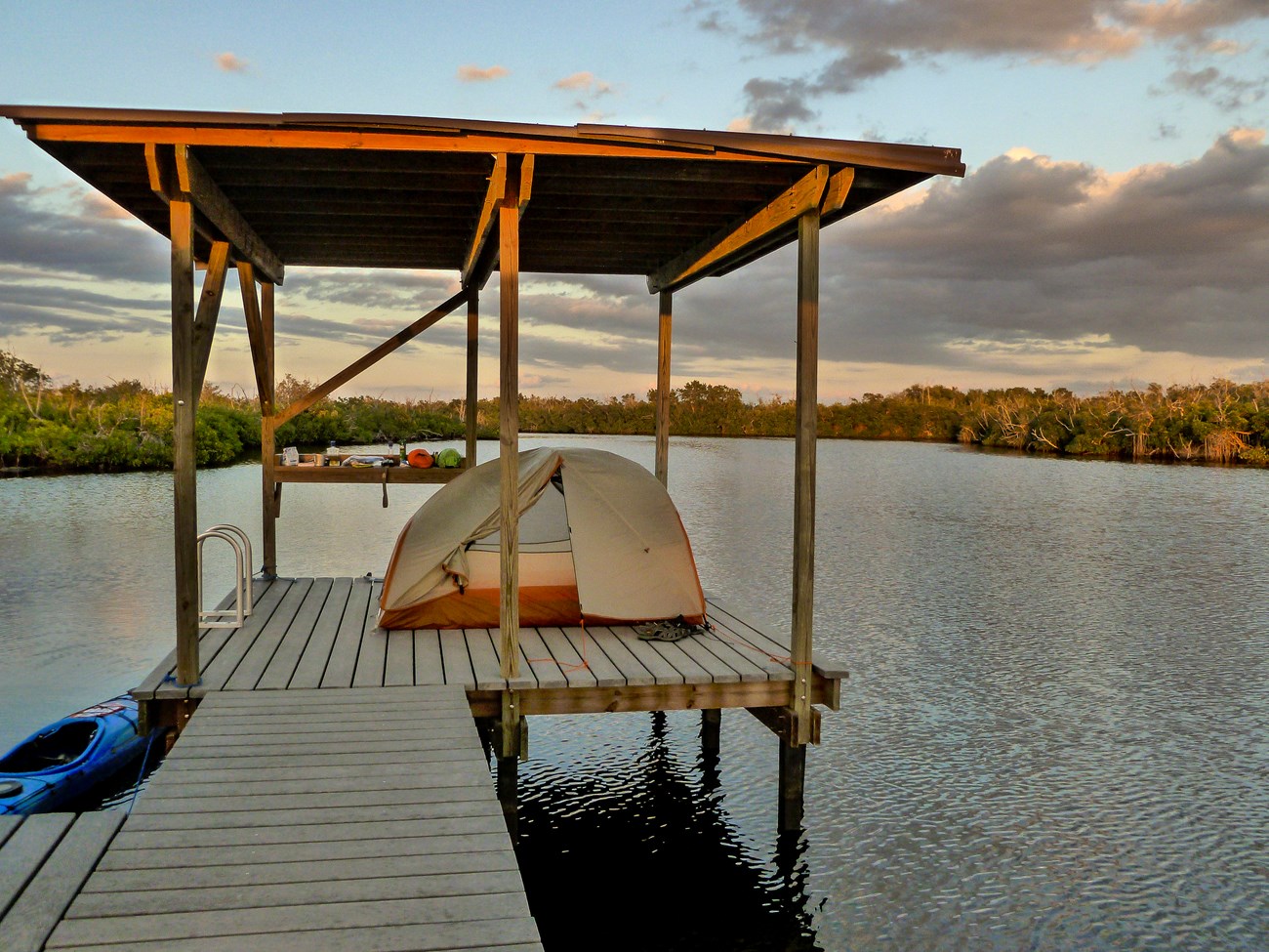 A tent and camping gear on a lifted wooden platform, above water. A kayak is on the side of the platform.