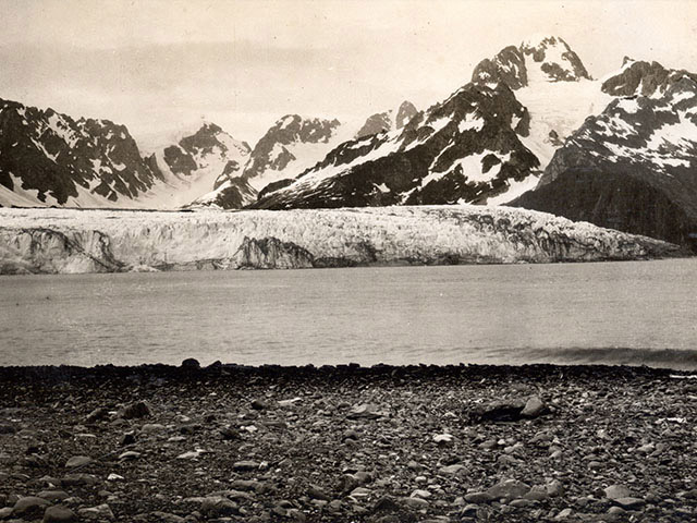 A black and white photo of a 1904 glacier coming down the mountains and forming a long wall on top of a lake. As the handle slides to the right, the image changes to a 2005 color photo of the same location, but there is no glacier on the lake. The glacier stops in the mountains before reaching the lake.