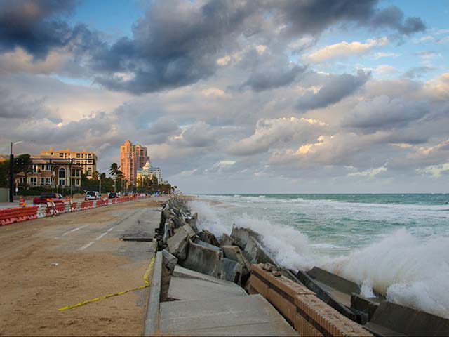 Ocean waves break away concrete sections of the road, while tall buildings line the opposite side.