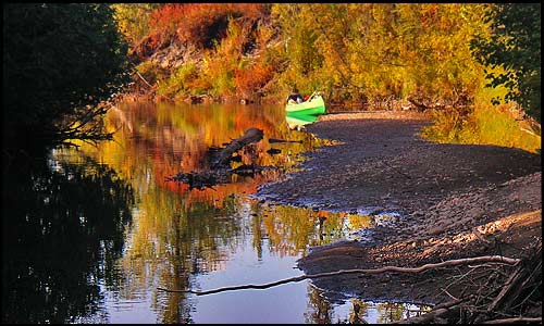 A green canoe sits along the shore of a small side stream
			that reflects the reds and golds of fall.
