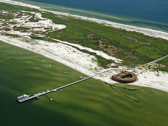 An aerial photo of a D-shaped brick fort on a long barrier island with blue-green water and white sandy beaches on both sides and green vegetation in the middle.