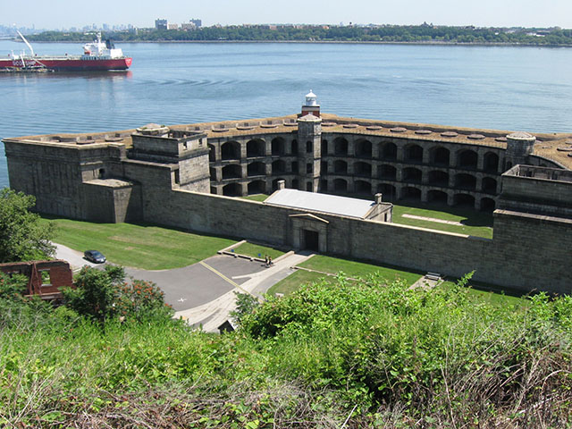 A three-story trapezoid-shaped fort looks over the harbor, and a cargo ship passes by. The land on the opposite side of the harbor is covered with buildings.