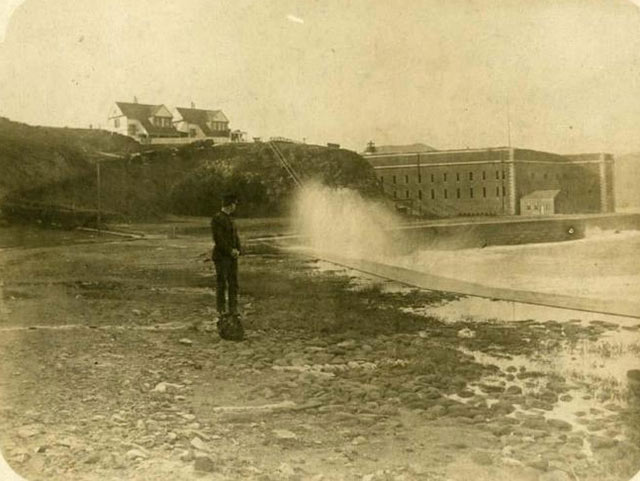 A black and white photograph of Fort Point. There is no bridge, but a building sits on the bluff behind the fort. A uniformed man stands on the walkway as salt-spray comes over the edge of the walkway. As the handle moves to the right, the image changes to a color photo of the brick fort, with the orange Golden Gate Bridge behind and above it. A ranger watches the salt spray from behind the wood-and-chain stanchions on the walkway. 