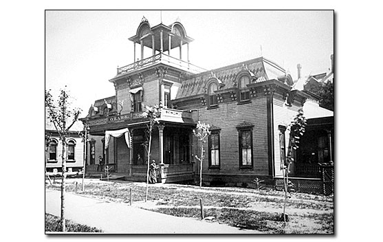 Black and white photo of the two-story Ozark Bathhouse, small leafless sapling trees are in front of the building next to the sidewalk. The building has a clapboard front and Mansard roof with a tower in the middle front with typical Victorian era ornamentation.