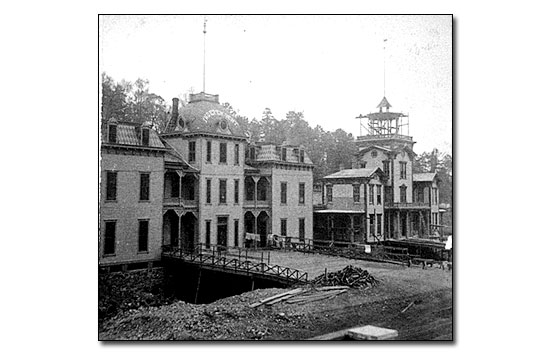 Black and white photo of two story structure, probably newly constructed because the foundation is exposed showing the depth of a basement and there is a pile of debris out front. A bridge is leads from the sidewalk to the door. There is a low railing along each side of the bridge and laundry is hanging on the railing near the entrance. The building consists of three bays connected by a veranda on first and second floors. The two end bays have a Mansard Roof and the center bay has a dome with the word MAURICE on it.