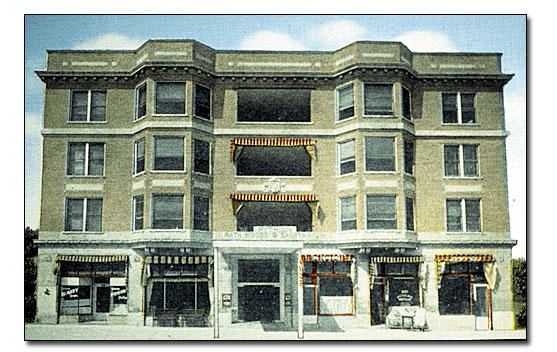 Tinted posted card of front of Pythian Sanitarium and Bathhouse in 1924. It is a four-story building with gray possibly limestone lower level and tan colored brick on the upper stories. There's an ornamentation of horizontal rectangles between the second and third stories and above the fourth story below the roof line. At that level there is also a dentition line directly above the windows. The windows and door of the street level are not symmetric while the widows on the upper levels are with a large central window, flanked by angled oriels, with a double paned window centered between the oriels and the end of the building. There is what looks like another storefront entrance at the far left end of the building because there is an inset doorway. There are red and yellow striped awnings that are pulled in on the street level windows and the second and third story central windows. 