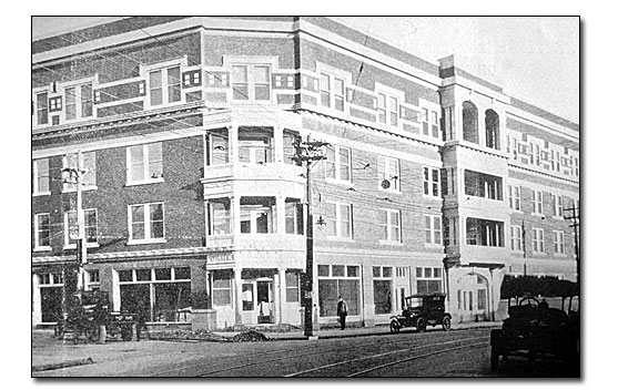 A black and white photo of the Woodman of the Union building. It is four stories tall, brick with light colored glazed terra cotta window surrounds and balconies. The building sits on the corner of two streets and the main doorway is on the corner. There are decorative balconies on the second and third stories above the entrance and at an entrance in the middle of the right or Malvern Avenue side with balconies above. There is a vehicle parked on both sides of the building and one in the foreground on the opposite side of the street. You can see the street car lines in front of the building and there are utility poles with service lines on the same side of the street as the Woodman building.