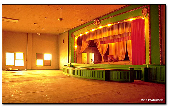 Colored photo. Large room with paper over floor to protect wooden floor, large stage on the right side. The front of the stage and all supporting columns and above the stage are painted bright green with decorative trim with gold leaf. The stage curtains are orange and red velvet. There are stage lights lining the sides and above the stage. DD Photoworks